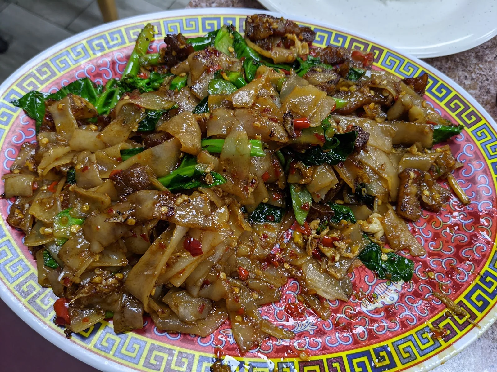 An oval pink-rimmed melamine plate of pad see ew with crispy pork, Chinese broccoli and a heavy scatter of red chili, on a speckled laminate table.