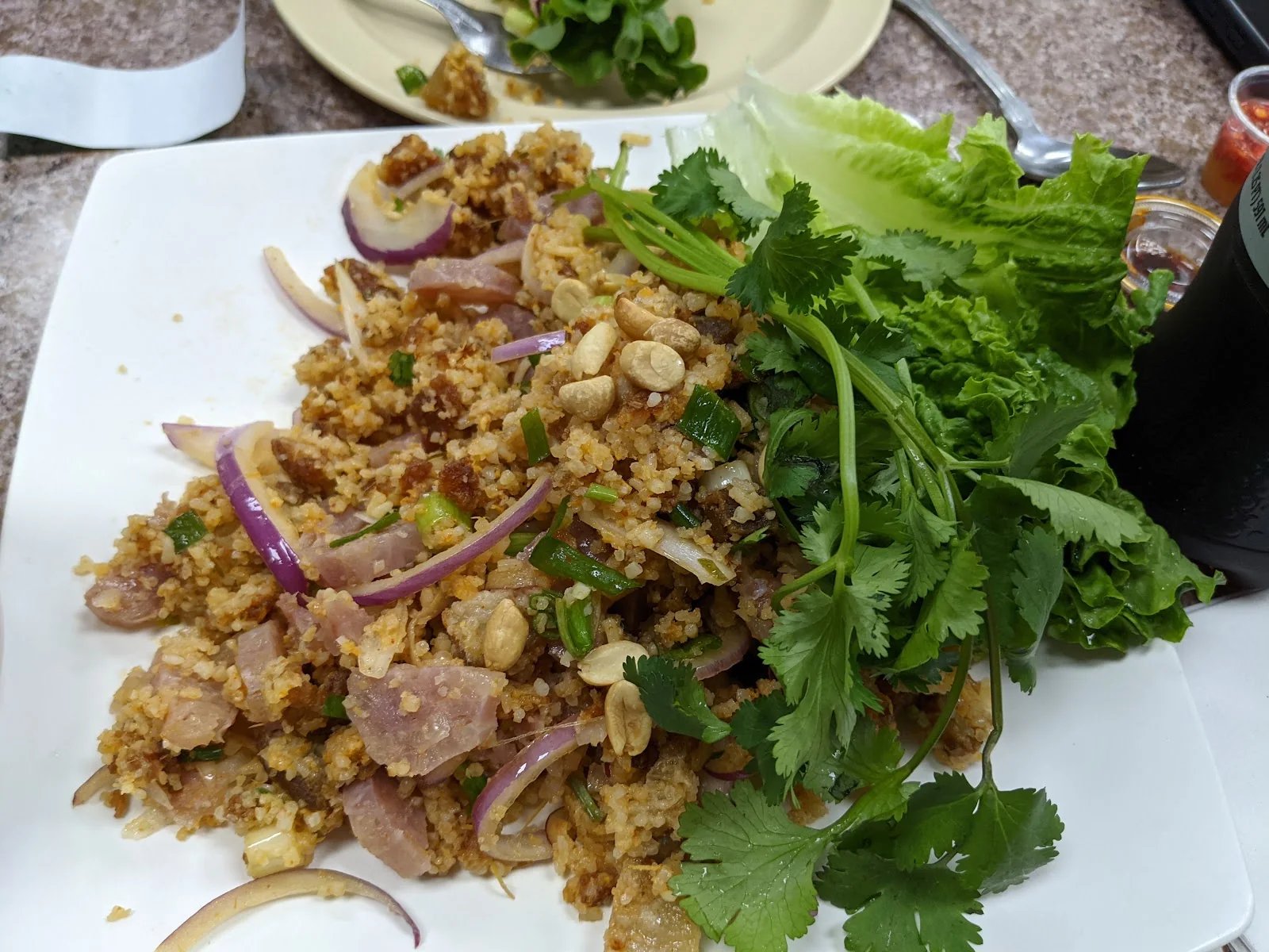 A white plate of crispy rice salad with pieces of sour sausage, sliced red onion, peanuts and a pile of fresh cilantro and lettuce leaves, on a speckled laminate table.