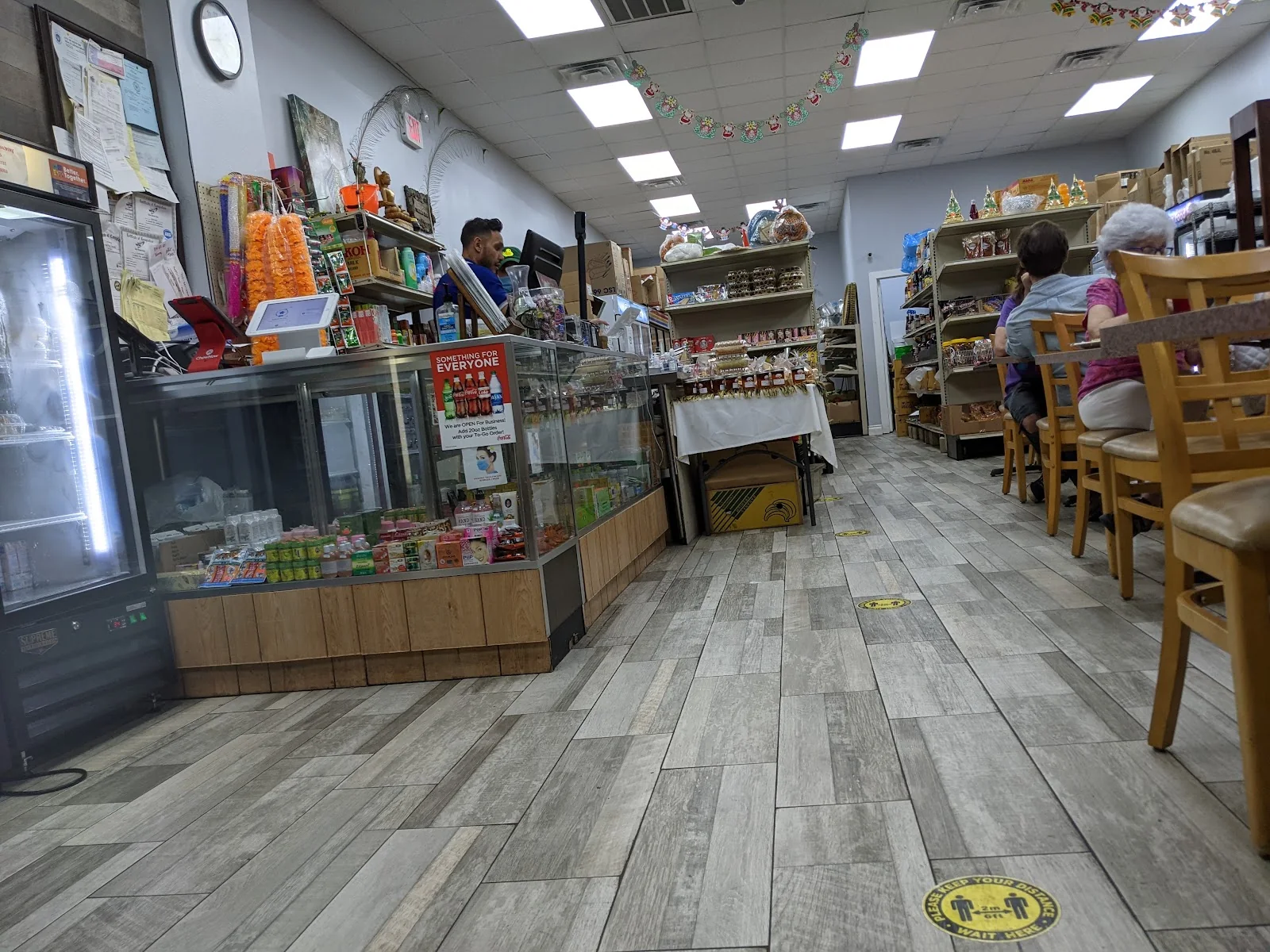 Inside a small grocery store, a glass deli counter and drink coolers on the left, shelves of packaged goods down the middle, and wooden restaurant chairs and tables at the back, on a grey wood-look tile floor.