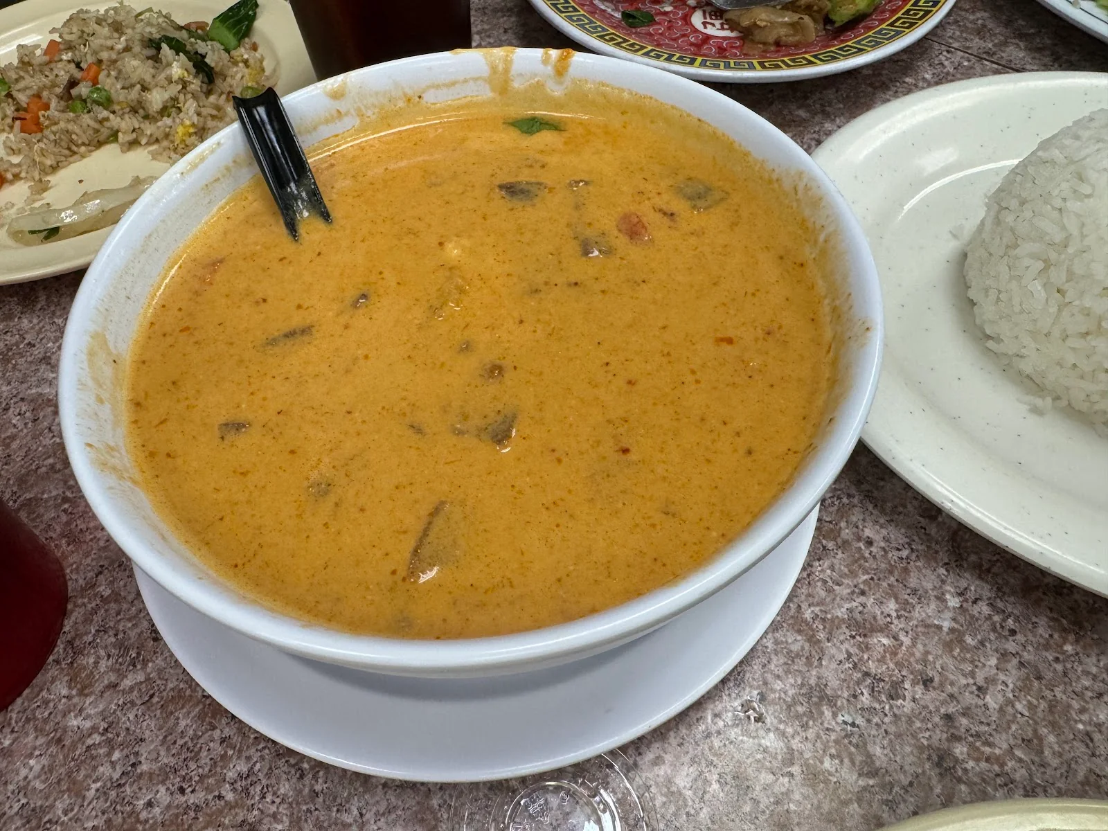 A white bowl of orange massaman curry with a black spoon resting in it, a plate of steamed rice to the right, the fried rice at the upper left and a pink-rimmed plate of noodles above, on a speckled laminate table.
