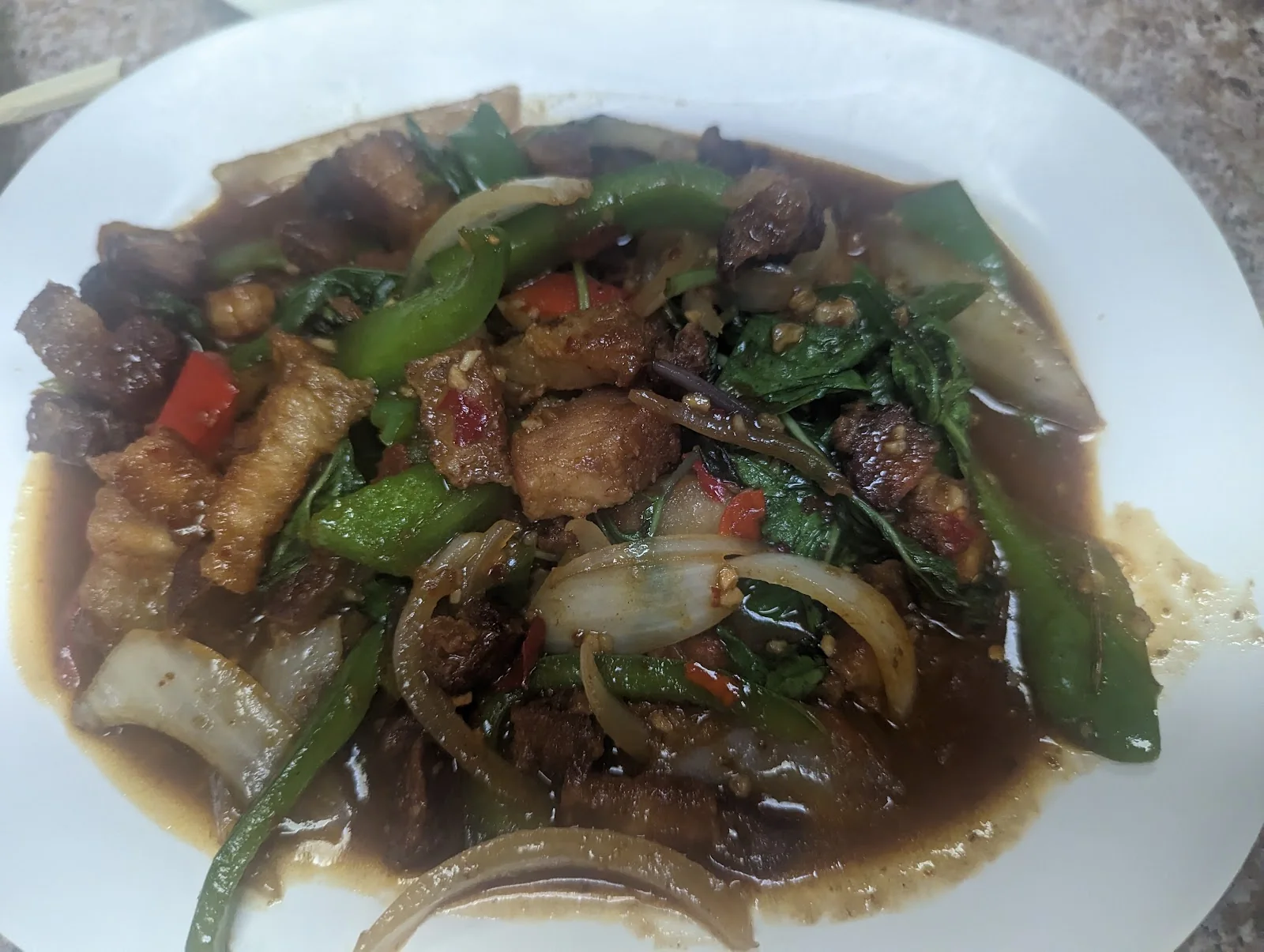 A white plate of stir-fried crispy pork belly with sliced onion, green and red bell pepper and basil in a dark chili sauce, on a speckled laminate table.
