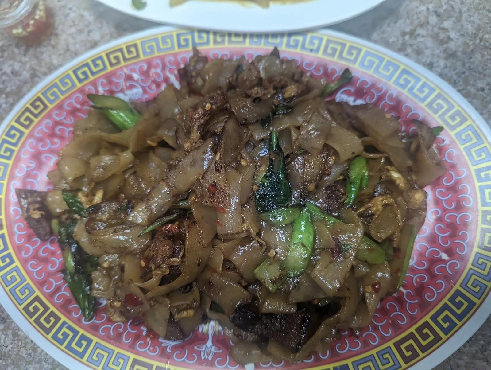 An oval pink-rimmed melamine plate of pad see ew, wide rice noodles with Chinese broccoli, egg and pieces of crispy pork belly, flecked with red chili, on a speckled laminate table.