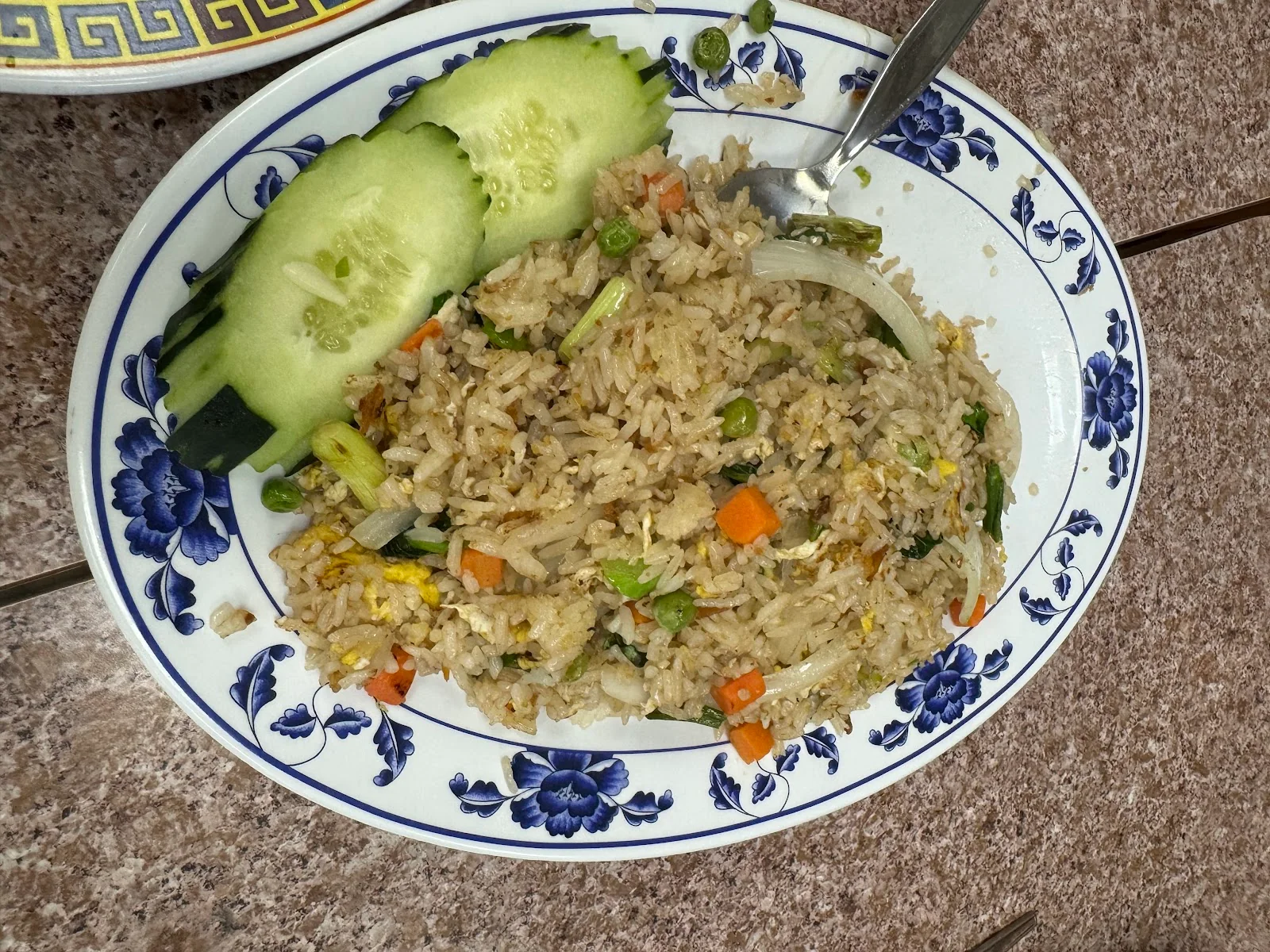 An oval blue-and-white patterned plate of fried rice with egg, peas and carrots, cucumber slices at the upper left and a spoon resting at the top, on a speckled laminate table.