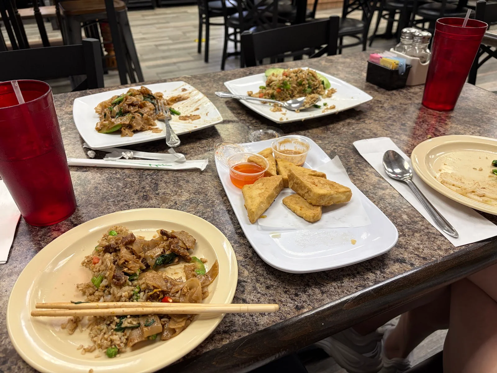 A restaurant table with rice and wide noodles on a round plate in front, chopsticks across its edge, fried tofu wedges and two dipping sauces in the middle, and square plates of noodles and fried rice behind, with red plastic tumblers and dark wooden chairs.