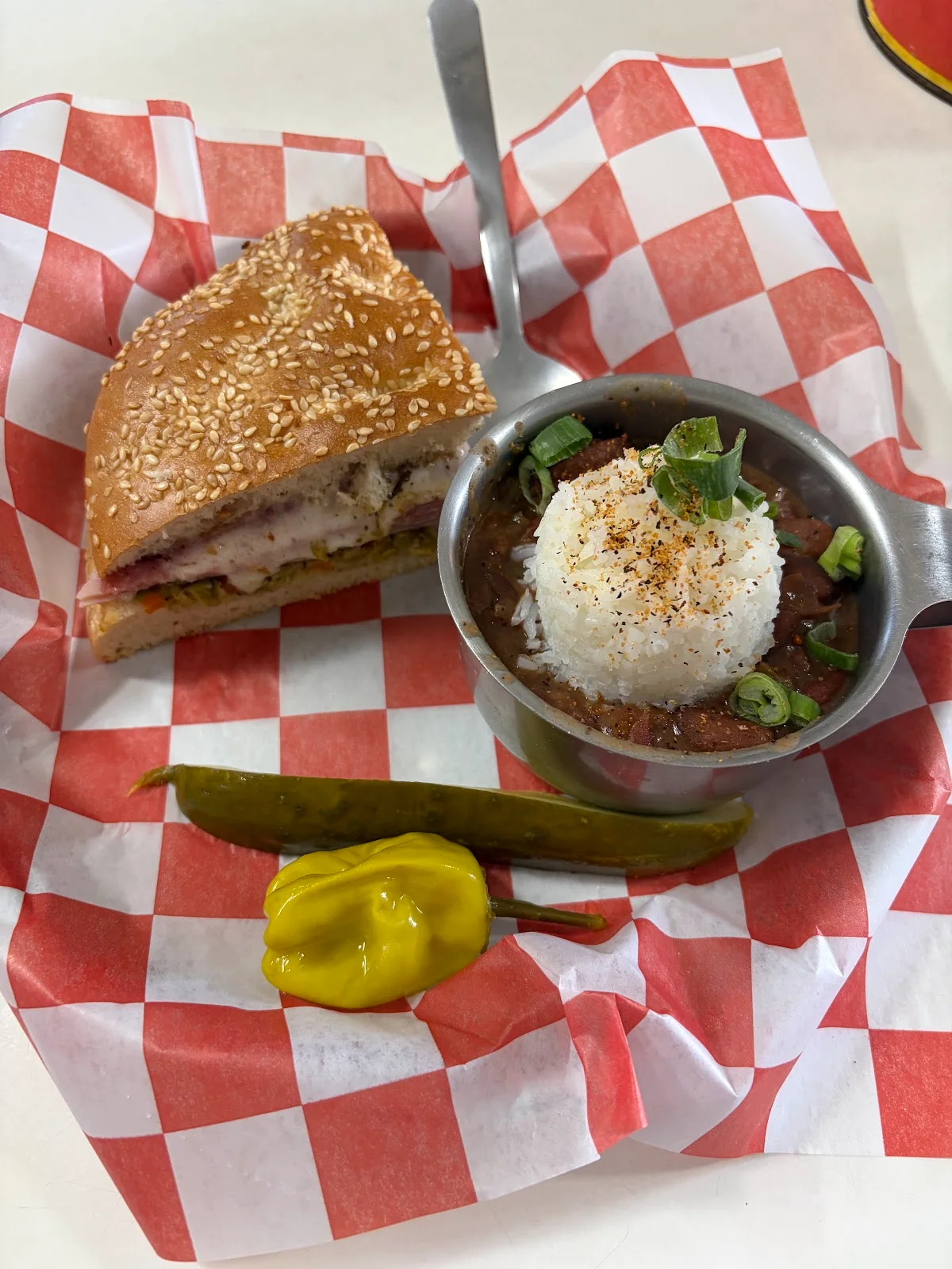 A quarter of a seeded muffaletta beside a small steel cup of red beans and rice topped with green onion, with a pickle spear and a pepperoncini, on red-and-white checked paper.