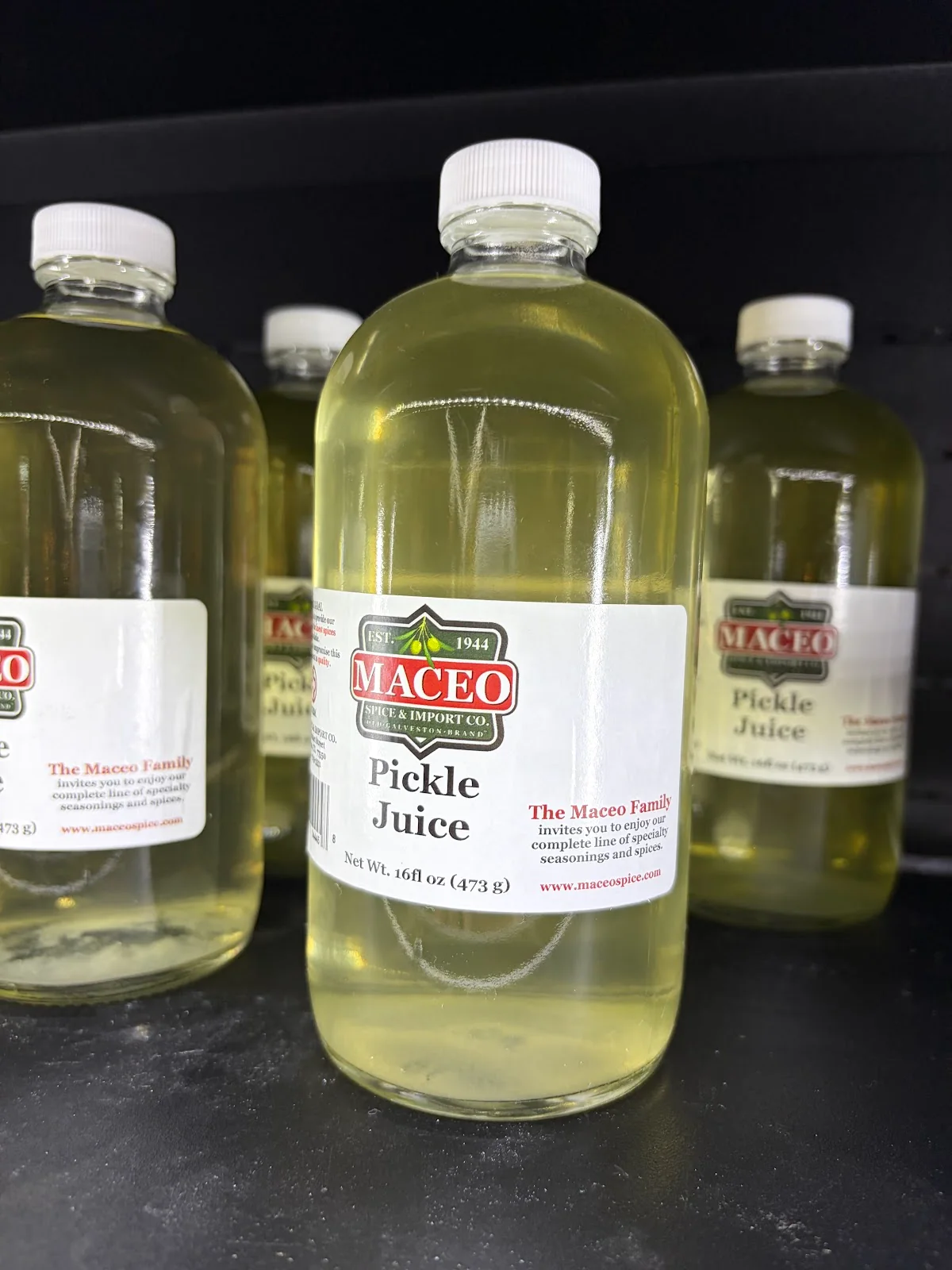Several glass bottles of Maceo Pickle Juice with white caps and red-and-green Maceo labels on a dark shelf.