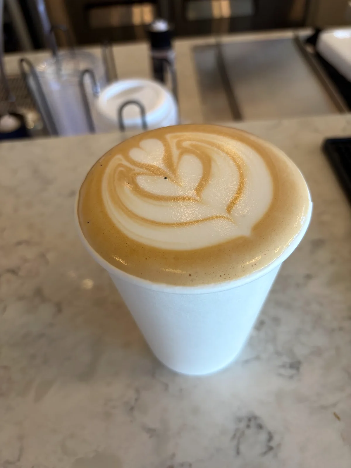 A latte with a heart of white foam in a plain white paper cup on a marble counter, cup lids and a syrup bottle out of focus behind it.
