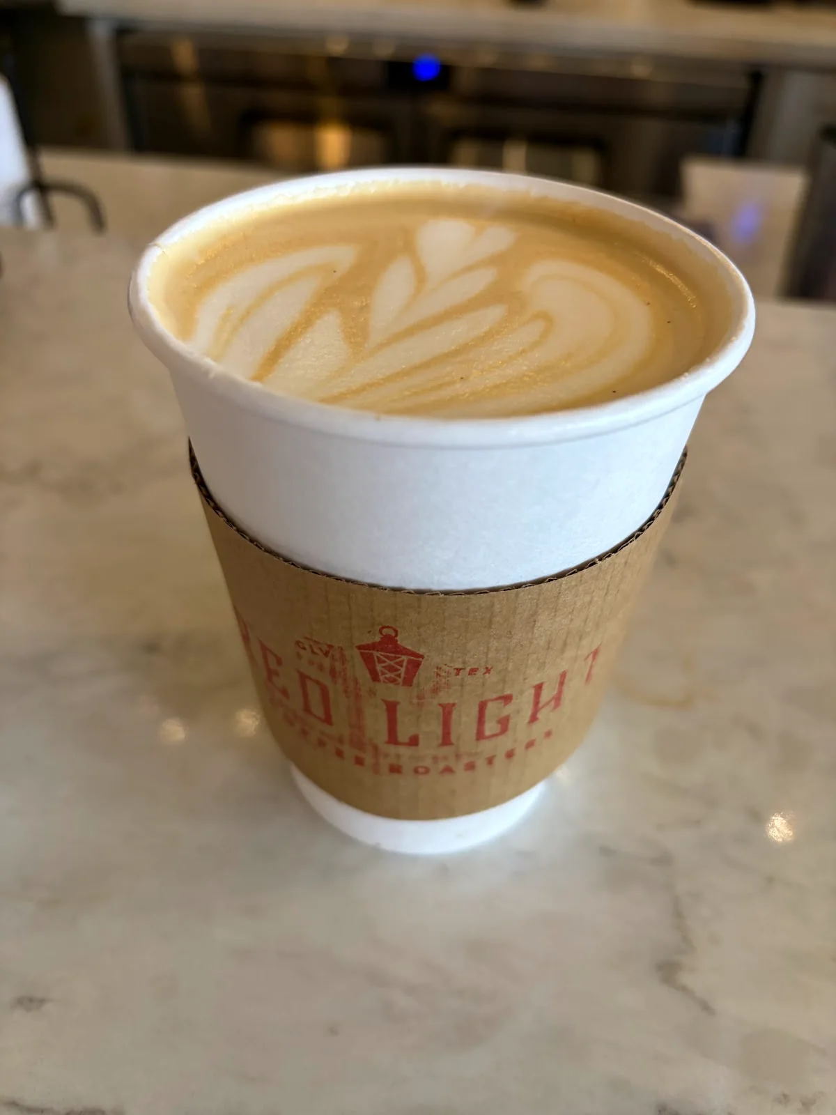A latte with a fern of white foam in a paper cup, its brown cardboard sleeve printed with the red Red Light Coffee Roasters lantern, on a marble counter.
