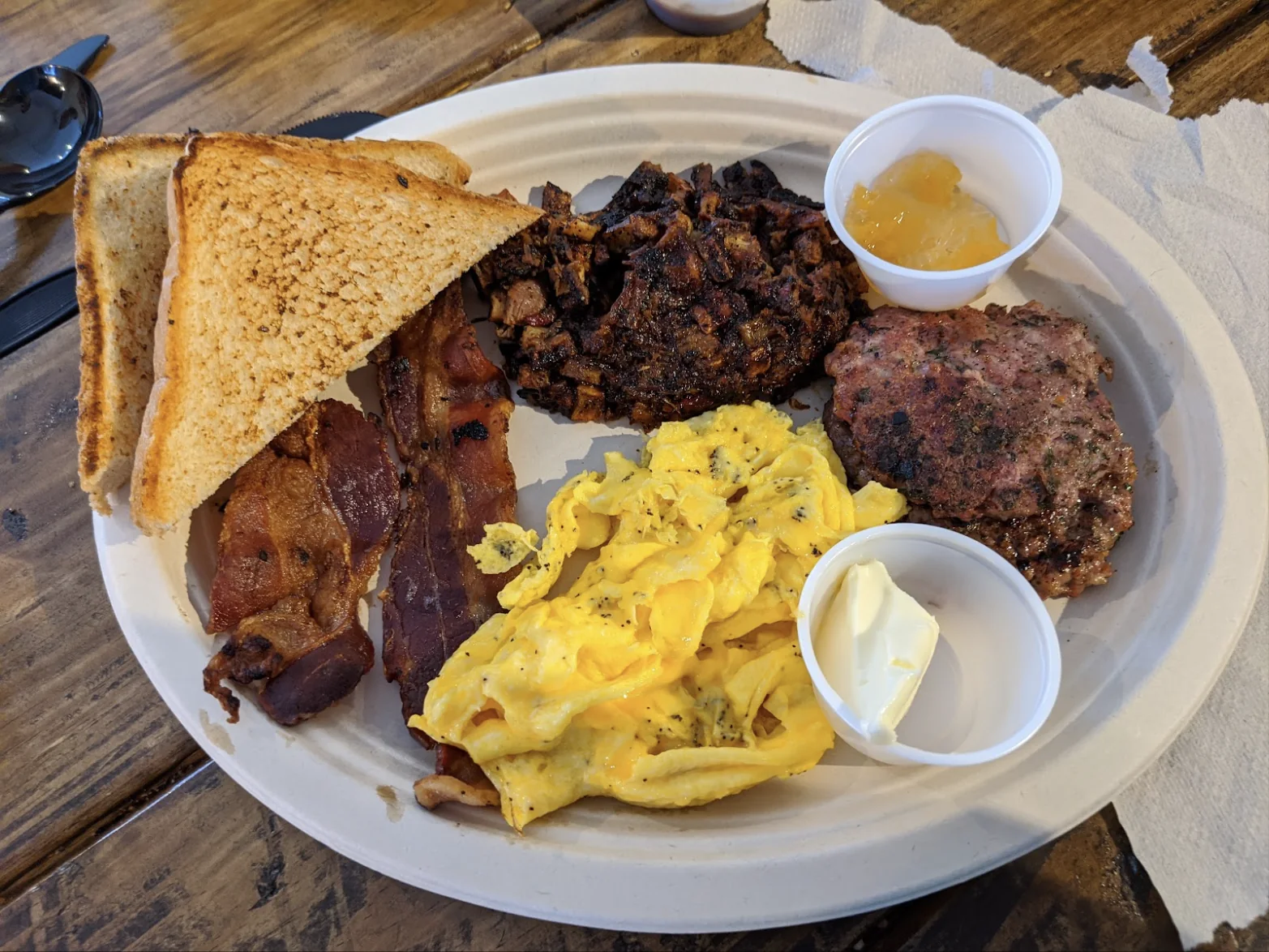 A paper plate with chopped brisket, scrambled eggs, bacon, a sausage patty, toast, and cups of butter and jam on a wooden table.