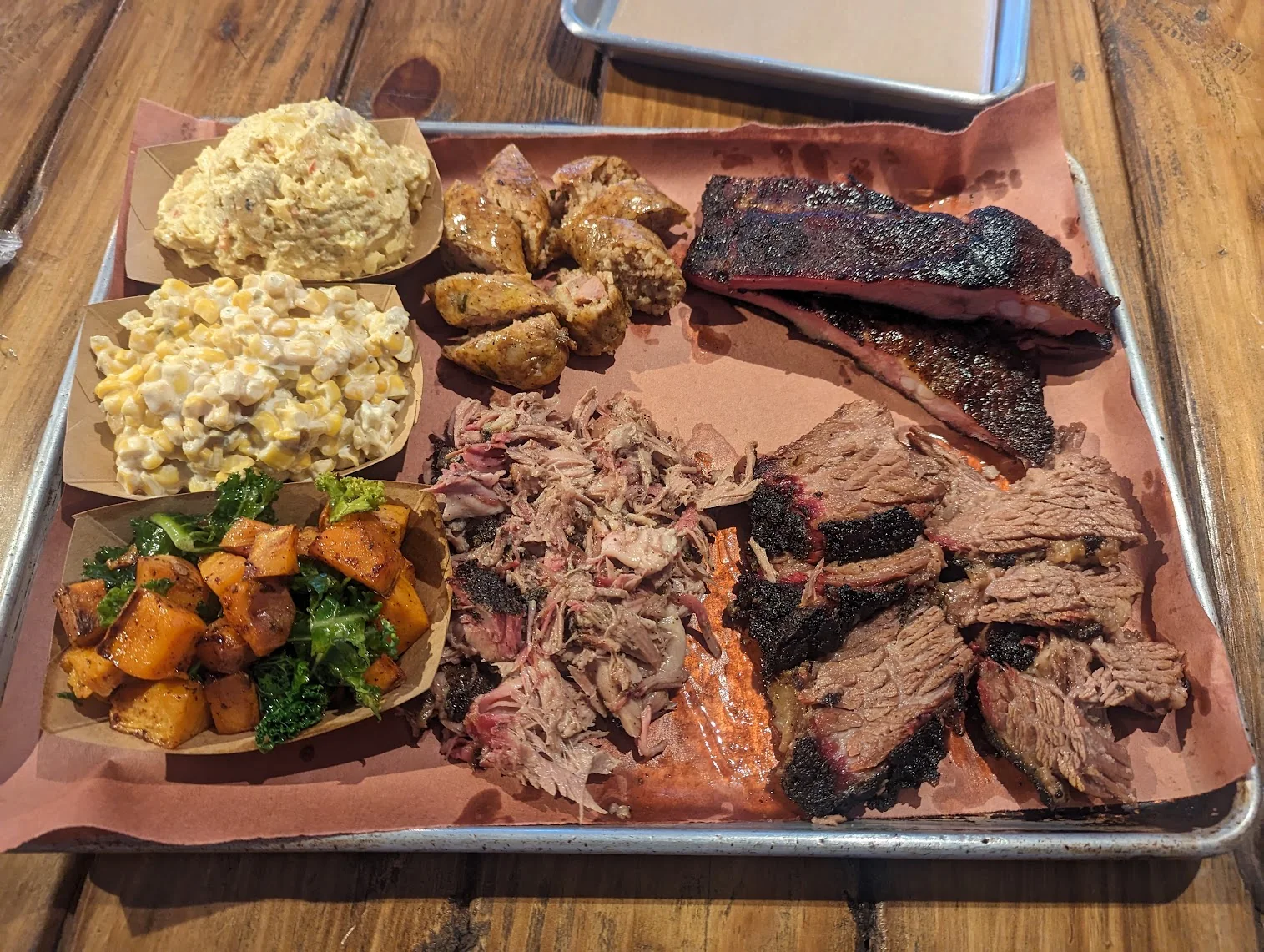 A butcher-paper-lined tray with sliced brisket in front, pulled pork, sliced sausage and pork ribs behind, and paper boats of potato salad, cream corn, and sweet potato and kale down the left side.