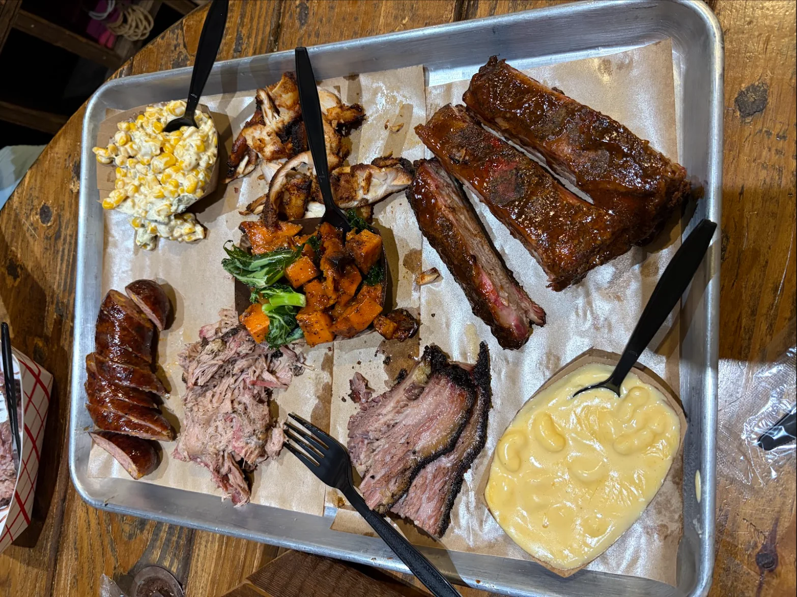 A sheet pan with sliced sausage, pulled pork, sliced brisket, pork ribs, cream corn, sweet potato and kale, and mac and cheese on butcher paper, on a wooden table.