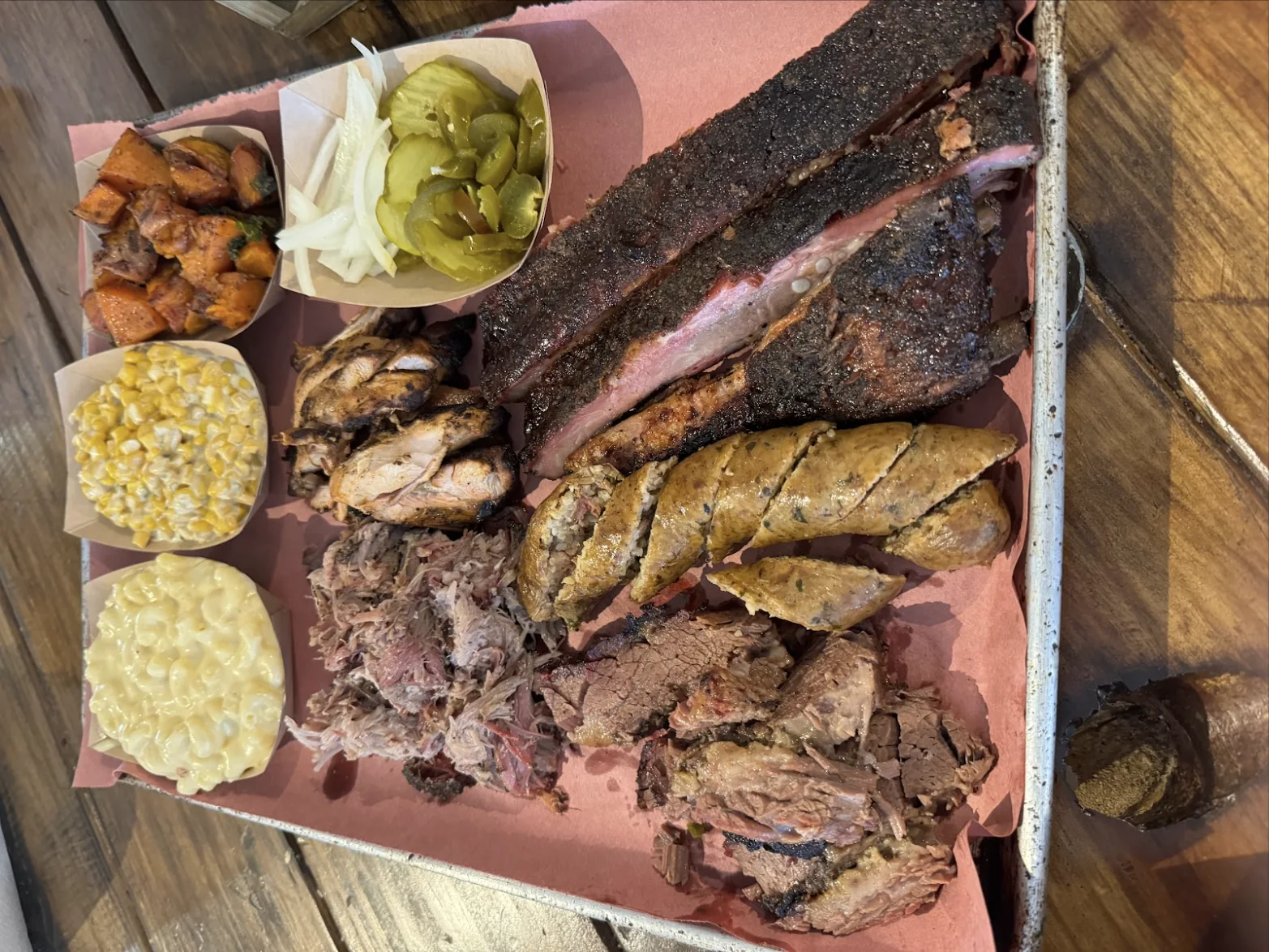 A butcher-paper-lined tray with sliced brisket, pulled pork, sliced sausage, pork ribs, smoked chicken, mac and cheese, cream corn, sweet potato and kale, and a paper boat of pickles and onions.