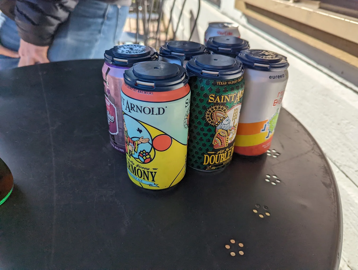 Six different craft beer cans, Saint Arnold and Eureka Heights among them, clipped together with a reusable carrier on a black metal patio table.